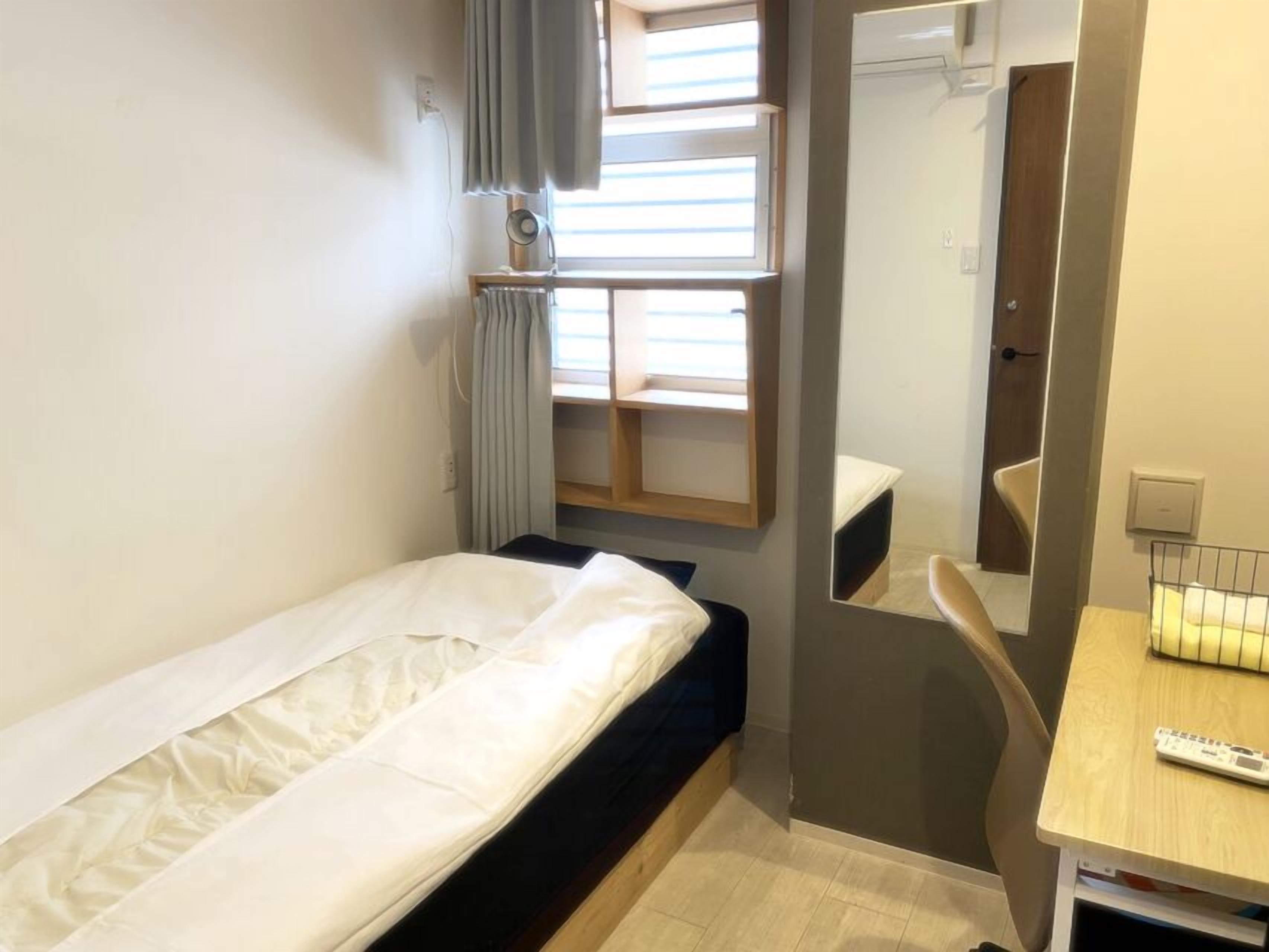Ein kleines, gemütliches Zimmer mit einem Einzelbett, einem Schreibtisch mit Stuhl, einem Spiegel und einem Fenster mit Vorhängen.