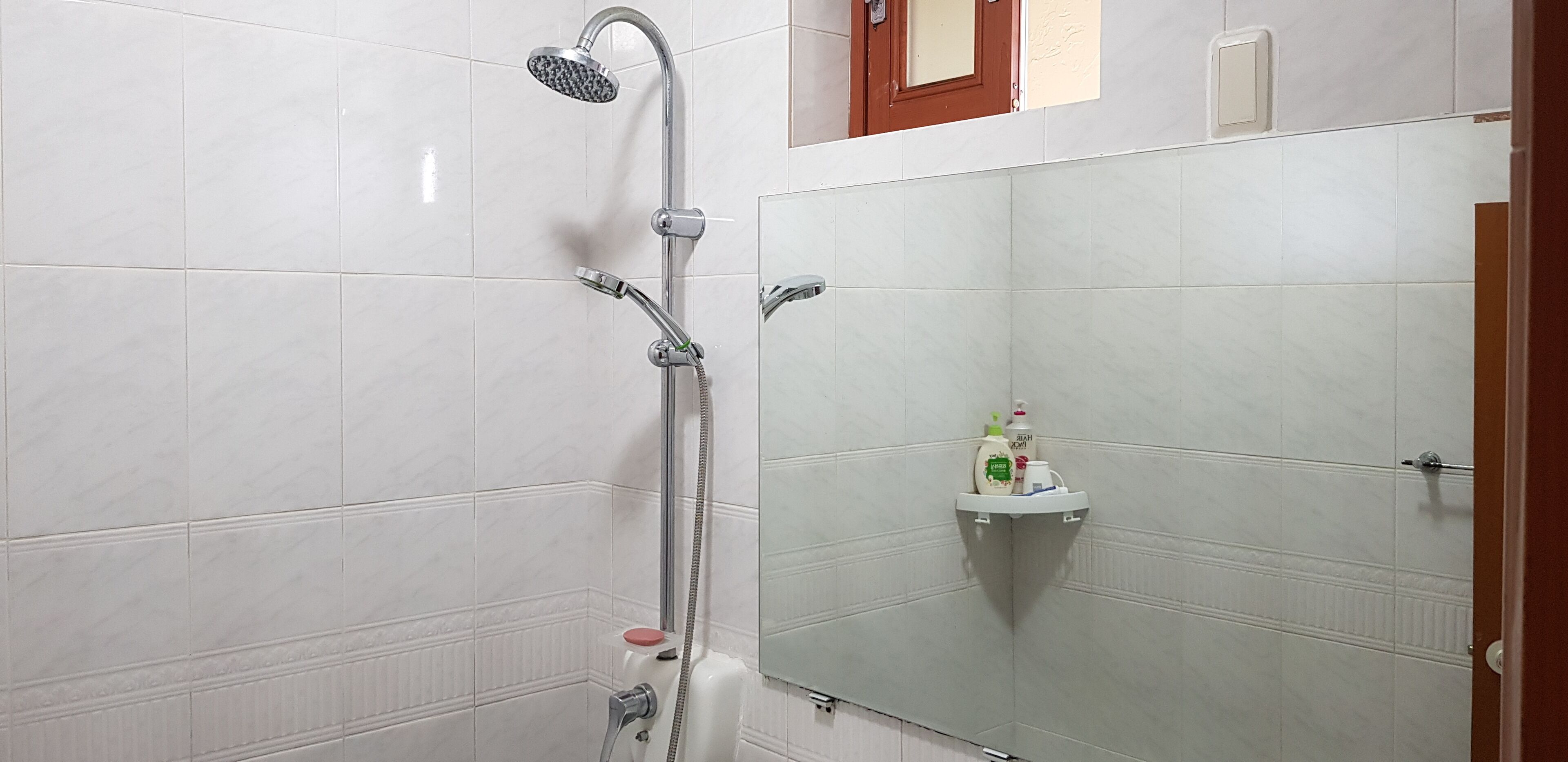 Un bagno con pareti piastrellate di bianco, una doccia con soffione manuale, un porta asciugamani e un dispenser di sapone su un piccolo ripiano.