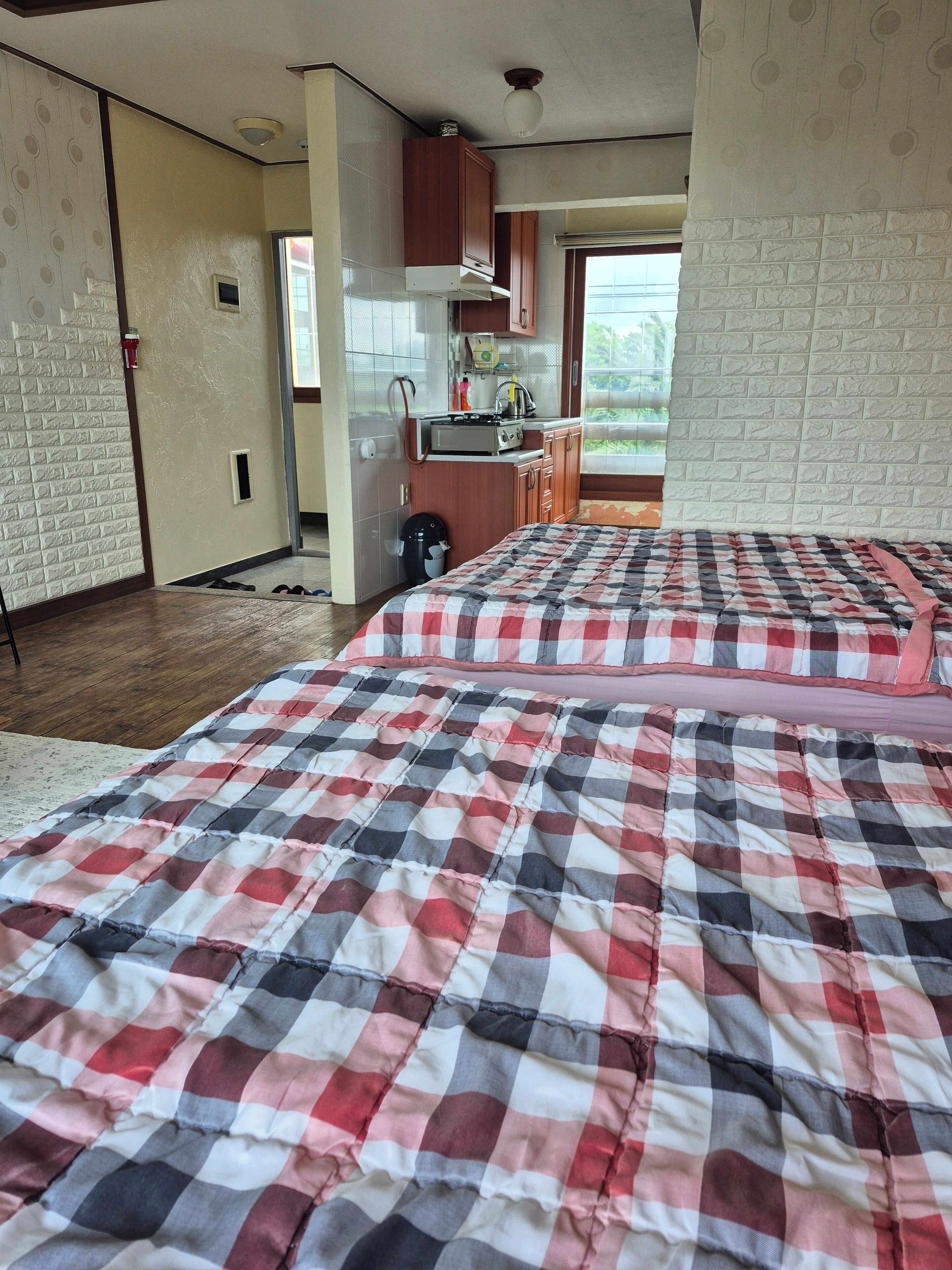 A bedroom with a checkered bedspread, a kitchenette with a stove, and a window with a view of greenery.