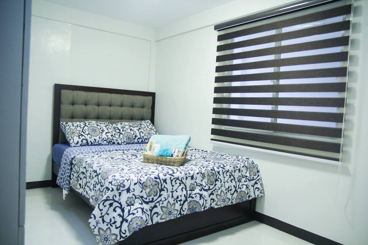 A bedroom with a bed, a headboard, a window with blinds, and a bedside table with a basket.