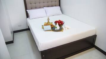 A neatly made bed with white bedding, a decorative pillow, a bowl of fruit, and a small sign on the bed.