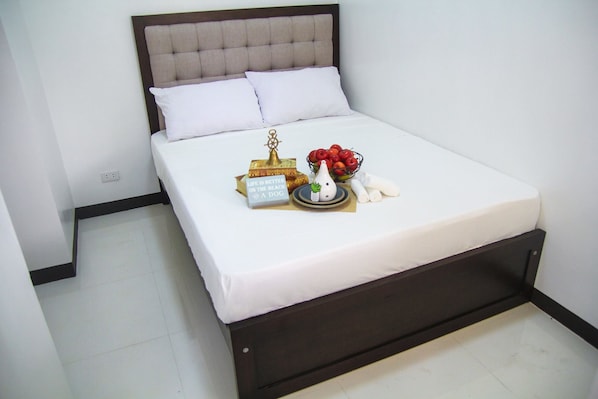 Una cama bien hecha con ropa de cama blanca, una almohada decorativa, un cuenco de fruta y una pequeña señal sobre la cama.