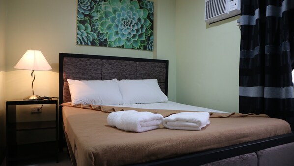 A neatly made bed with white pillows and a brown blanket, a bedside lamp, a wall-mounted air conditioner, and a large succulent plant. - Doniti Suites and Events Place (Mandaluyong)