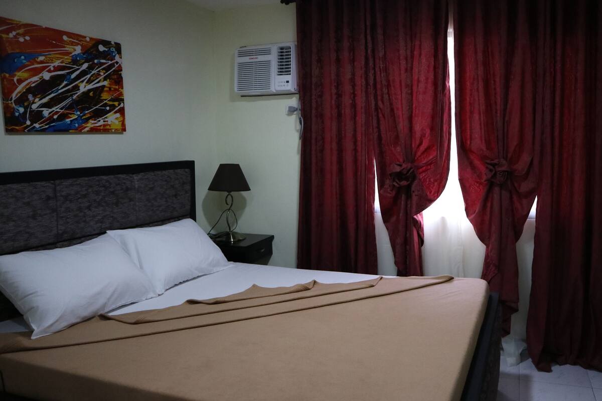 A hotel room with a bed, a nightstand, a lamp, a red curtain, and a wall-mounted air conditioner.