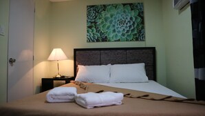 A neatly made bed with white linens and pillows, a bedside lamp, and a wall-mounted artwork of green plants. - Doniti Suites and Events Place (Mandaluyong)