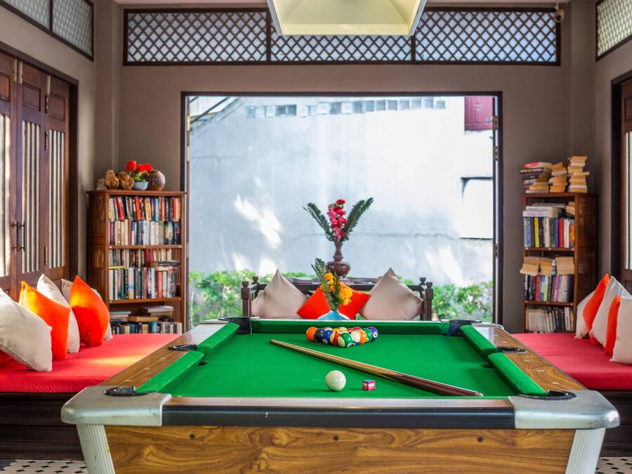 A pool table in a room with a sofa, bookshelves, and a view through a window.