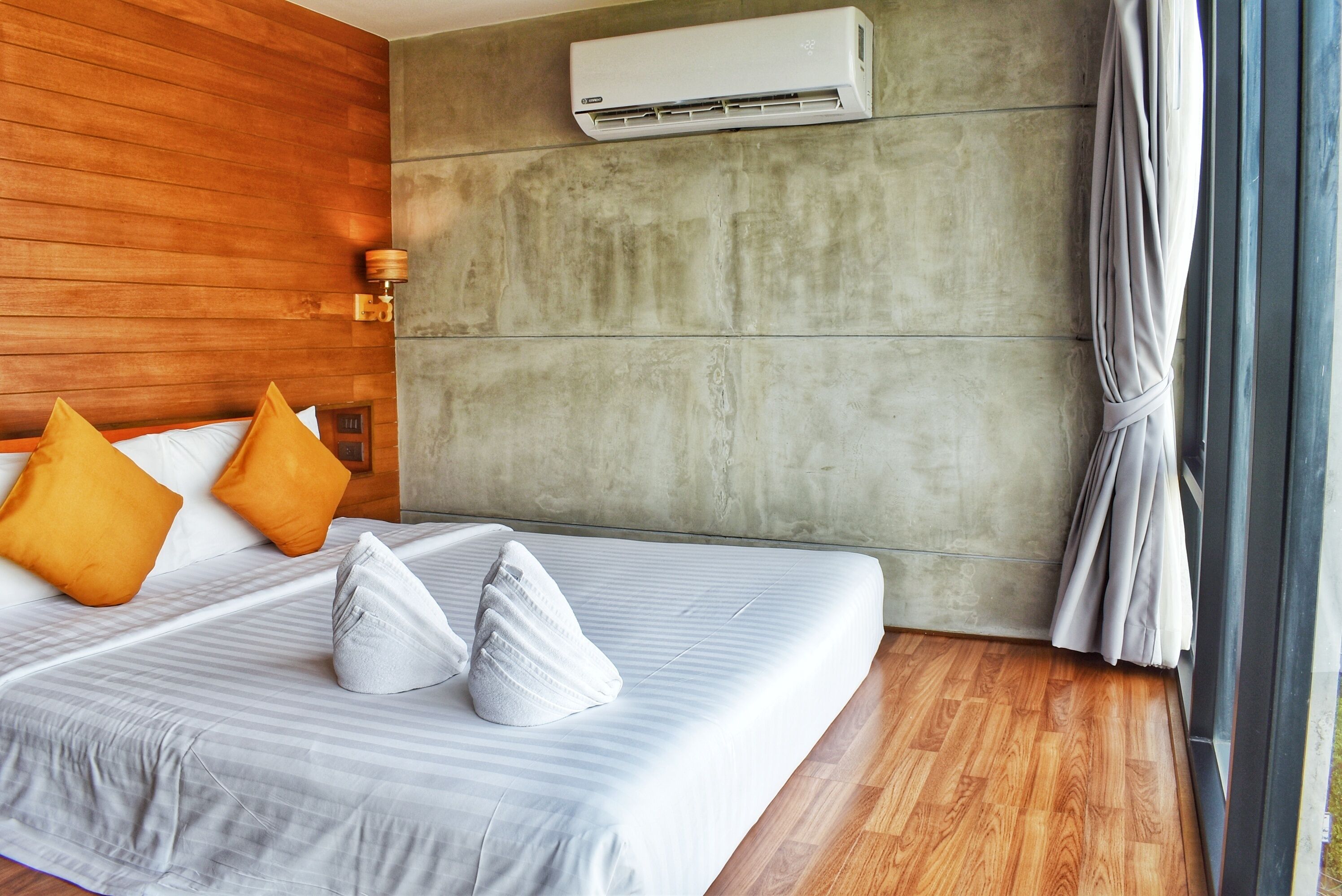 A hotel room with a wooden headboard, a large bed with white and gray bedding, two orange pillows, a gray pillow, and an air conditioning unit on the wall.