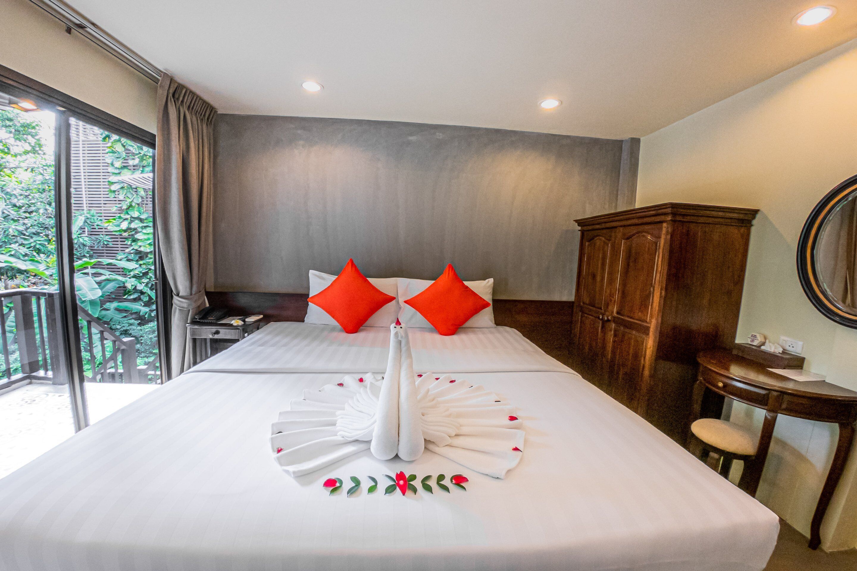 Habitación de hotel con una cama grande, dos almohadas rojas, un armario de madera, una mesita pequeña y un ventanal con vistas a zonas verdes.