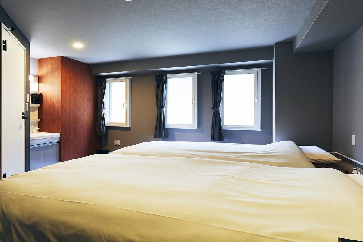 Habitación de hotel con dos camas, un armario de madera, una mesita pequeña y dos ventanas con cortinas.