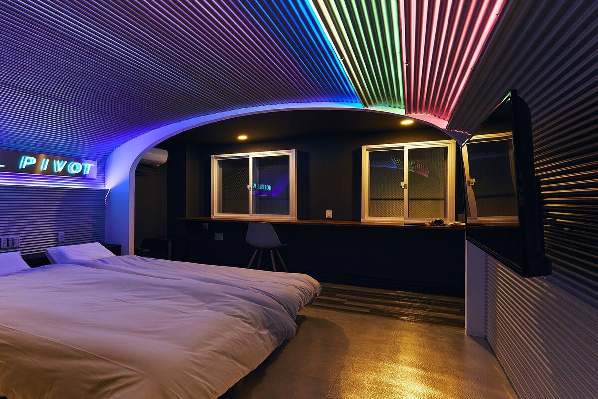 Una habitación de hotel con cama, escritorio y ventana. La habitación tiene un diseño moderno con techo curvo y un letrero de neón que dice "HOTEL PIVOT".