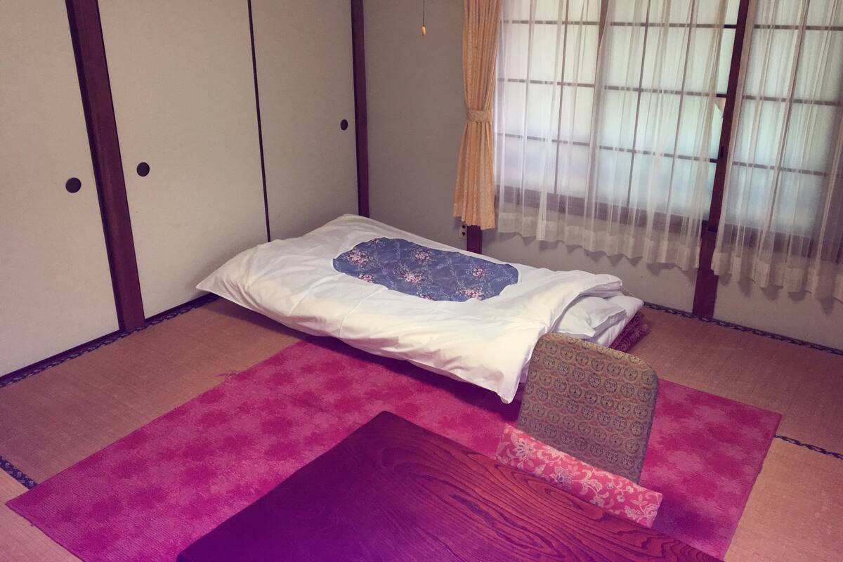 A room with a bed, a wooden bench, and a window with sheer curtains.