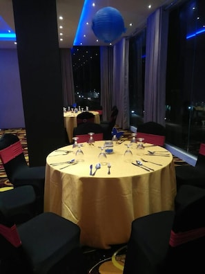 Uma sala de conferências com uma mesa redonda posta para uma reunião, rodeada por cadeiras com fitas vermelhas, e uma janela grande oferecendo vista da cidade à noite.