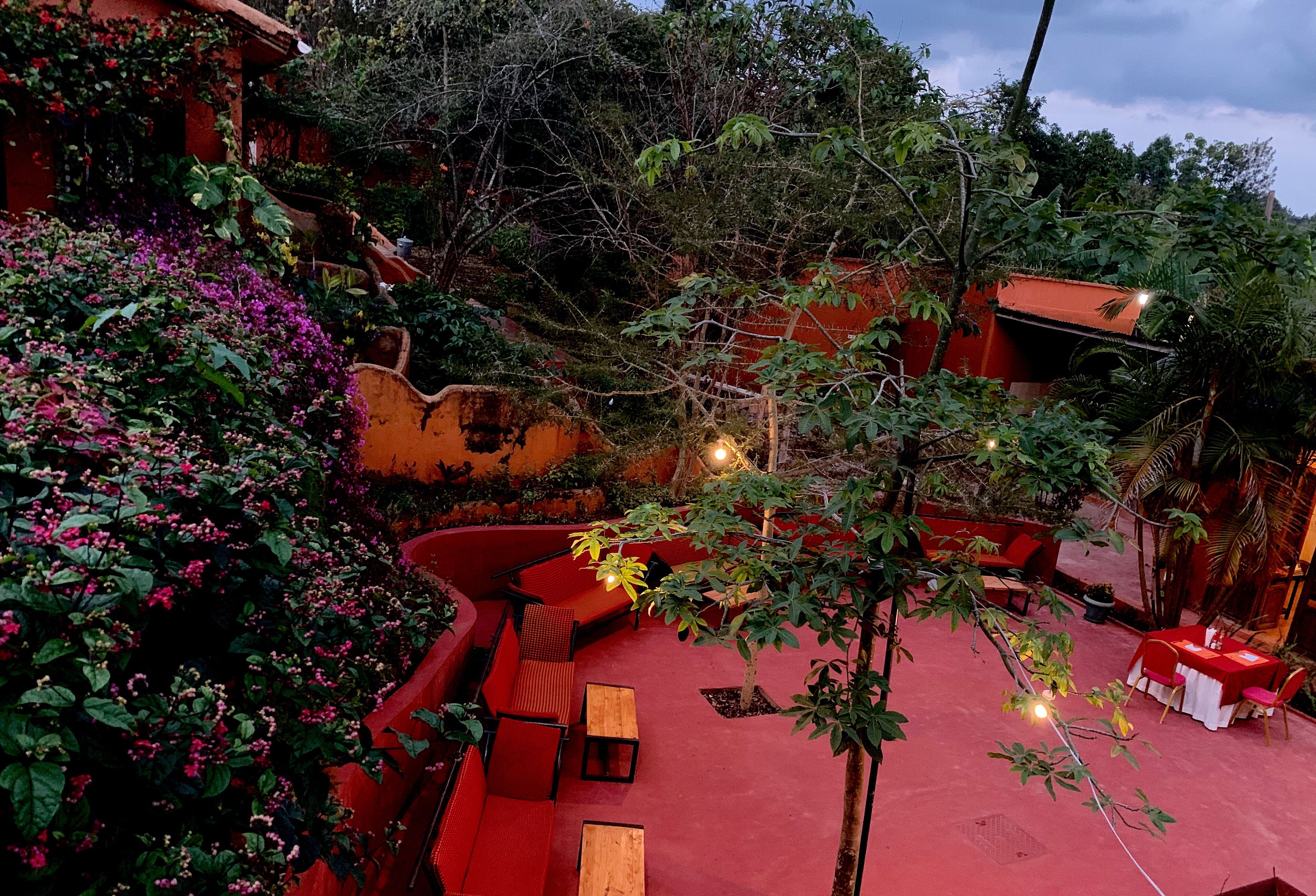 Un área de estar al aire libre con techos rojos, mesas y sillas de madera, rodeada de exuberante follaje y plantas con flores.