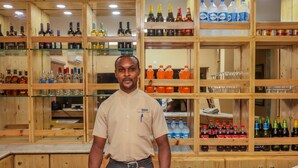 Un homme se tient dans un coin bar, devant des étagères remplies de bouteilles et de verres de toutes sortes.