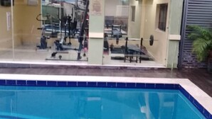 Une piscine dont le bord est recouvert de carreaux bleus, entourée d’un bâtiment doté d’une porte en verre donnant accès à une salle de sport.