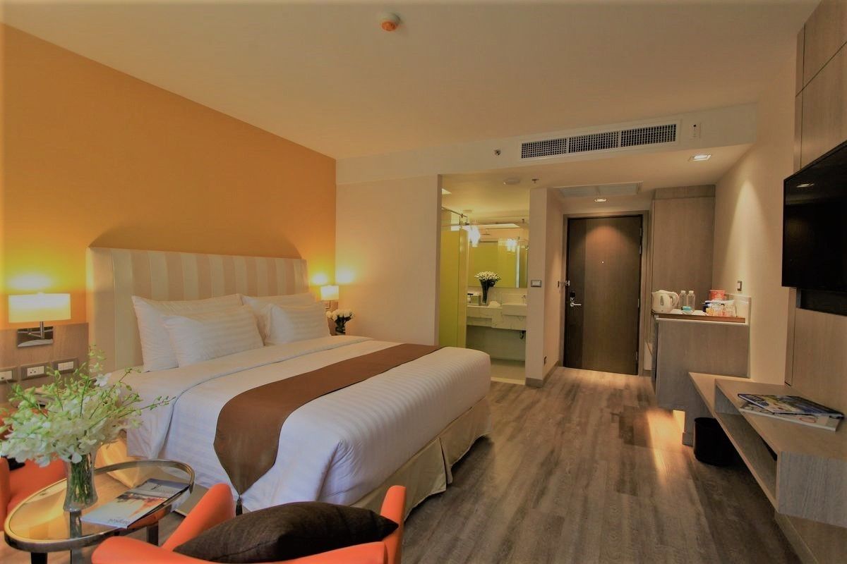 Habitación de hotel con una cama grande, un escritorio, una silla, una mesita con un florero y un televisor en la pared.
