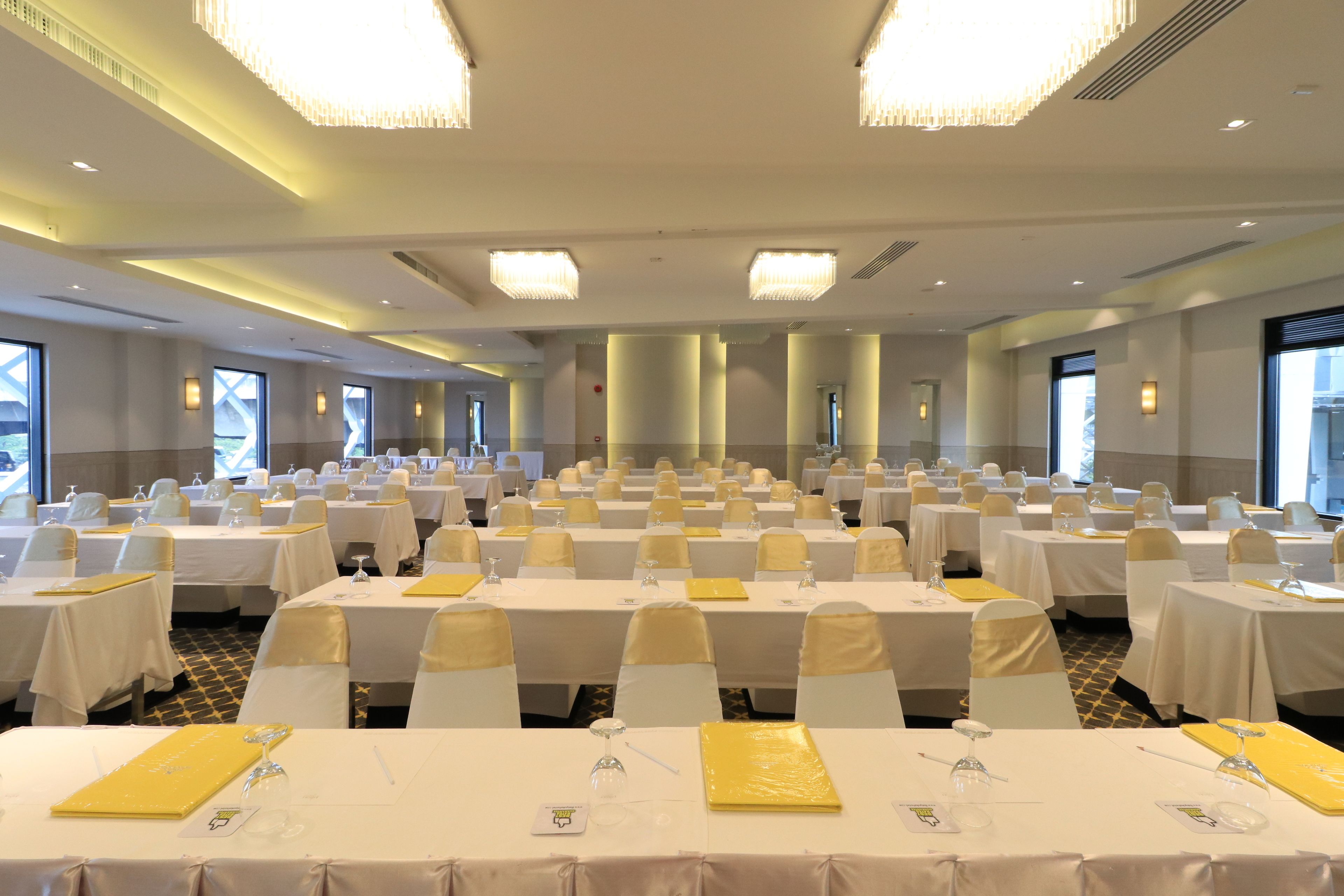 Un salón de banquetes con varias mesas dispuestas para eventos, con manteles blancos, cintas amarillas y cristalería.