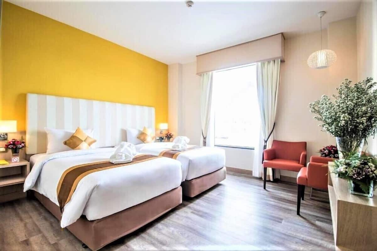 A hotel room with two beds, a red armchair, a large window with curtains, and a vase of flowers.