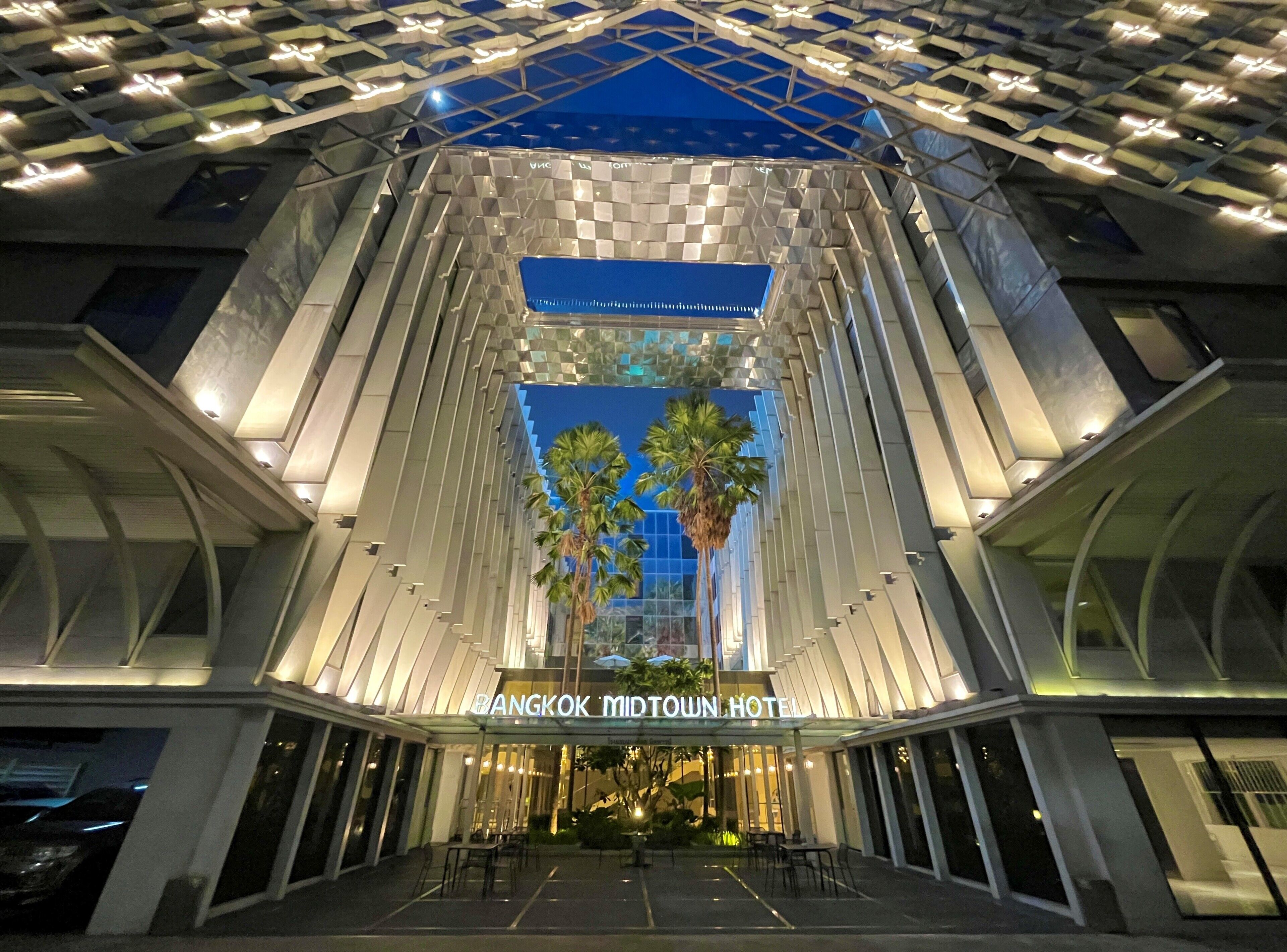 Hotel Bangkok Midtown, con un diseño arquitectónico moderno, que presenta una entrada destacada y una palmera en el centro.