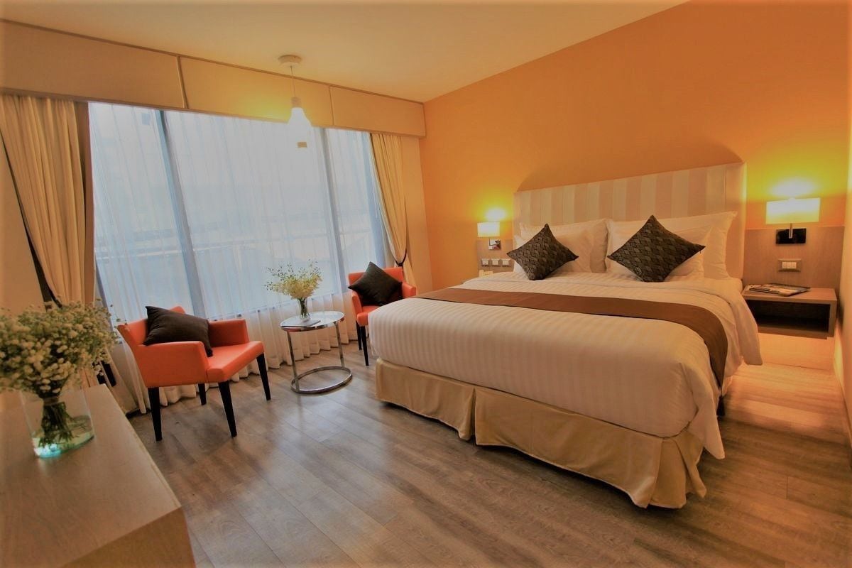 A hotel room with a large bed, two bedside tables, a small round table, a sofa, and a window with curtains.