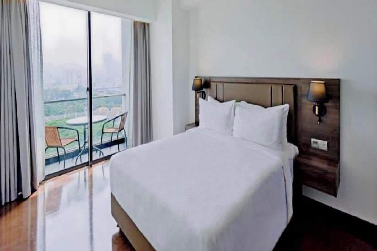 A hotel room with a large bed, a view of the city through a balcony, and a small table with chairs.