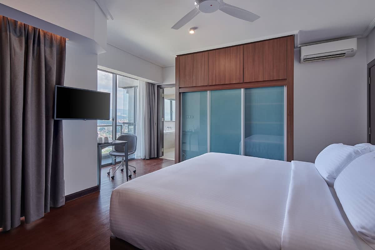 A hotel room with a large bed, a desk with a chair, a television, and a sliding glass door leading to a balcony.