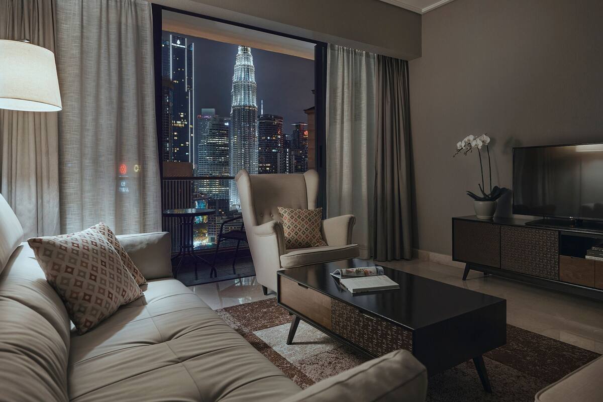 A modern living room with a sofa, armchairs, a coffee table, and a TV. A large window offers a view of a city skyline at night.
