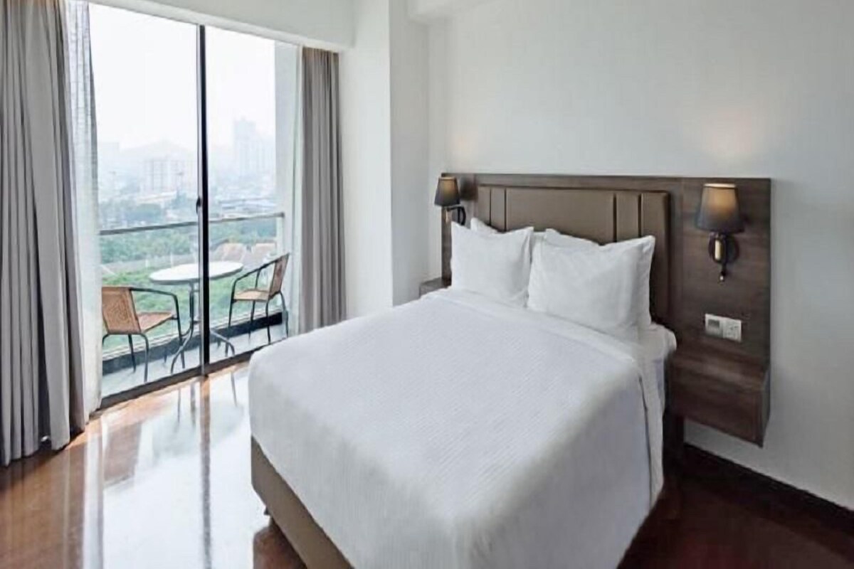 A hotel room with a large bed, a nightstand with lamps, a view of a cityscape through a sliding door, and a balcony with chairs.