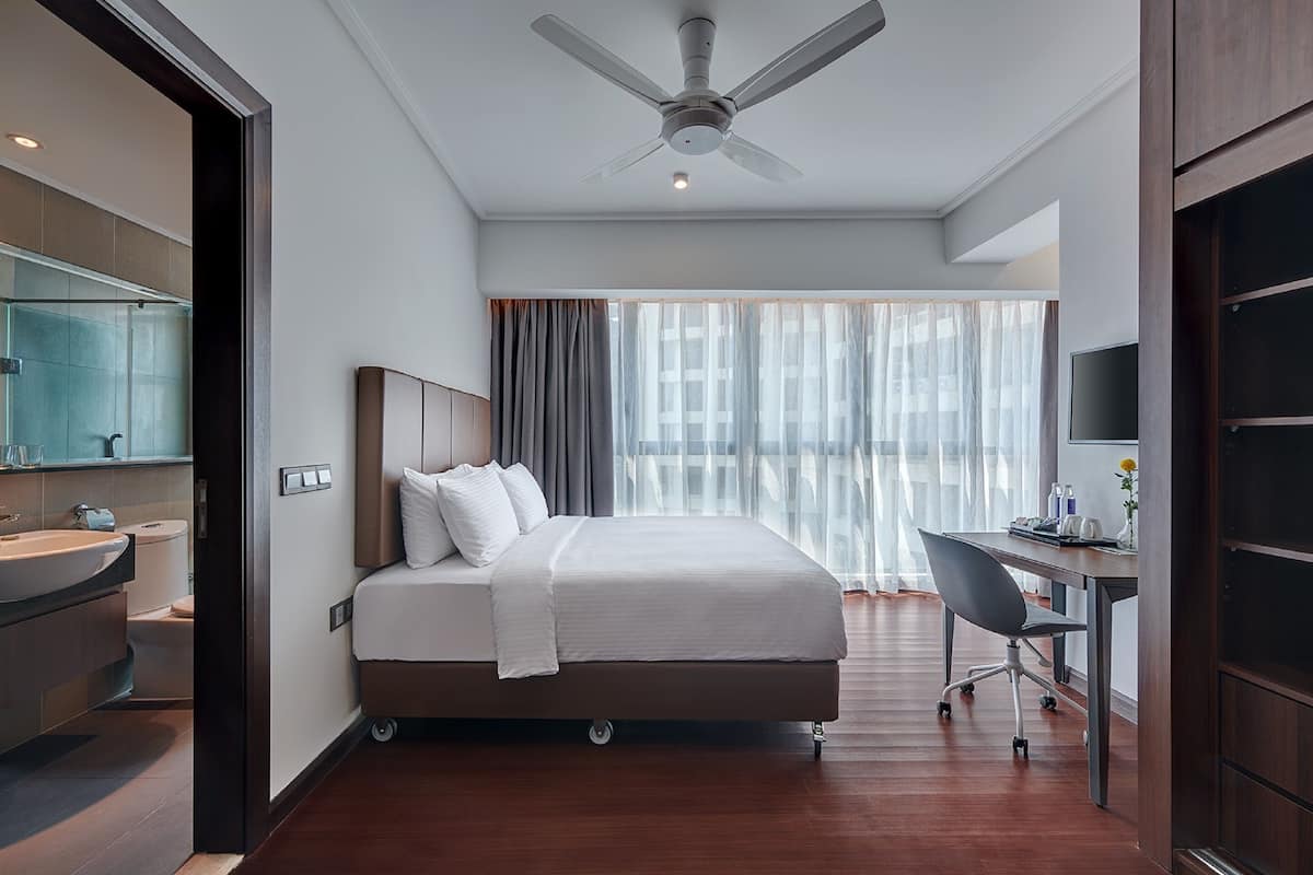 A modern hotel room with a large bed, a desk, a chair, a ceiling fan, and a bathroom with a sink and toilet.