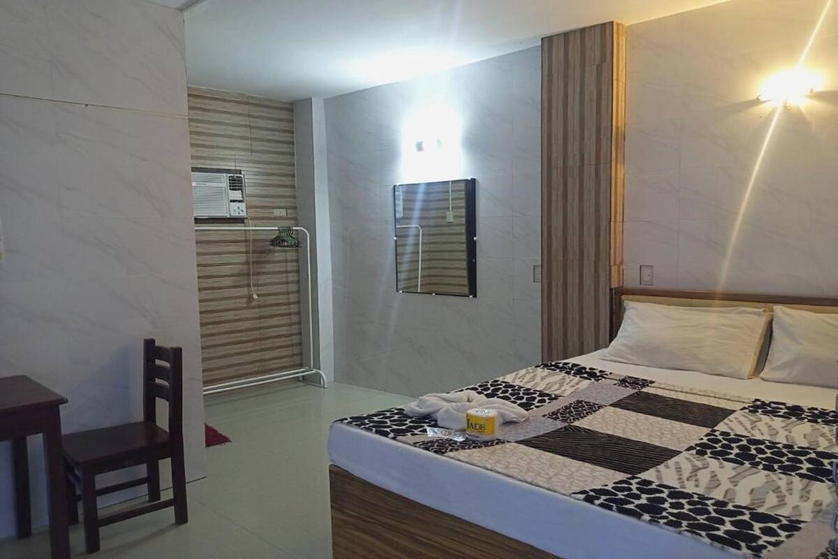 A hotel room with a bed, a desk, a chair, a wall-mounted mirror, and an air conditioning unit.