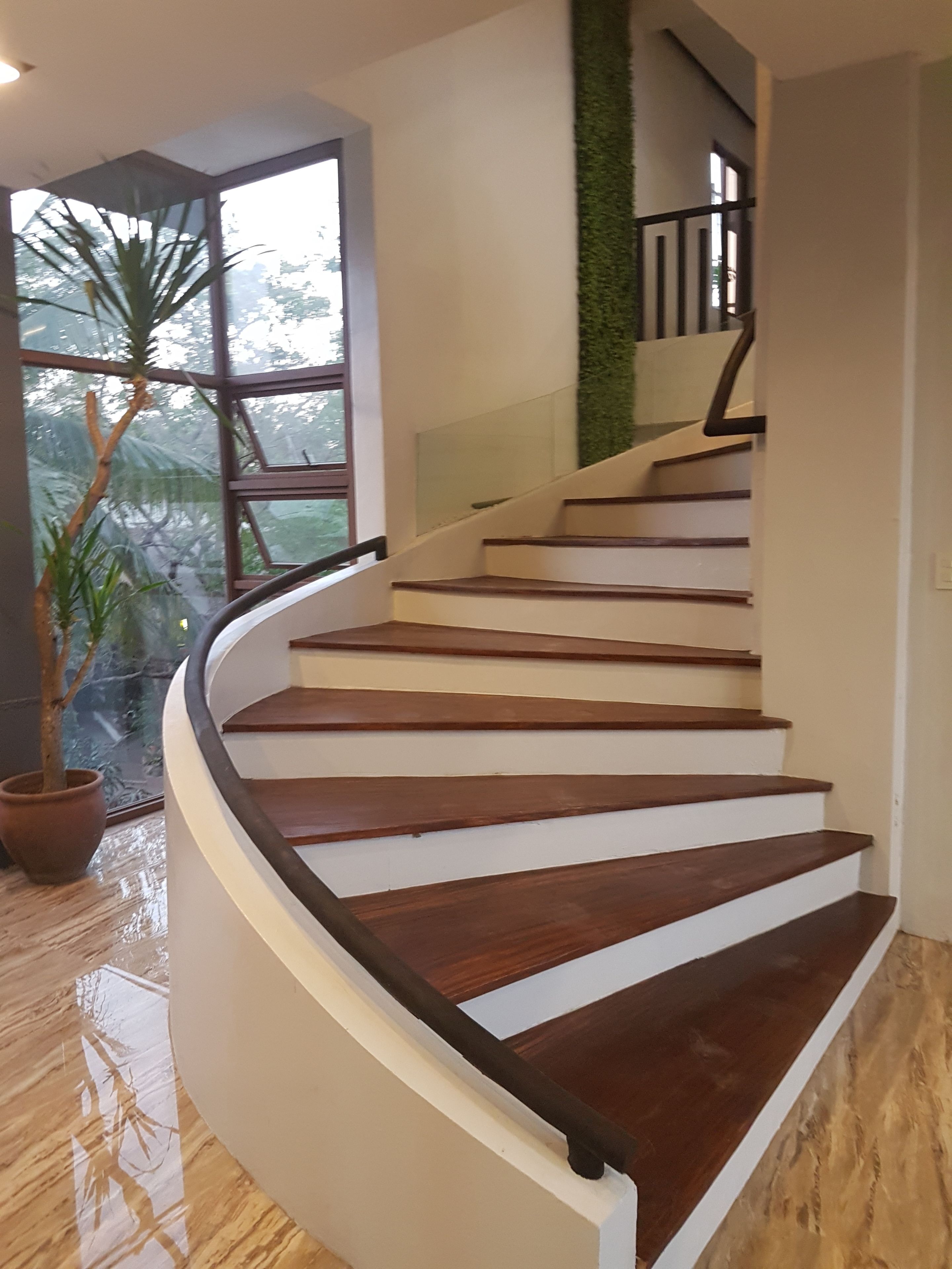 A spiral staircase with wooden steps and a black handrail, leading to a landing with a glass railing and a view of greenery outside.