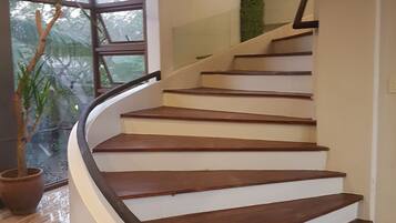 A spiral staircase with wooden steps and a black handrail, leading to a landing with a glass railing and a view of greenery outside.