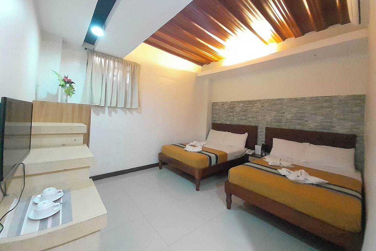 Habitación de hotel con dos camas, una mesita pequeña con una taza y platillo, y un televisor sobre un mueble.