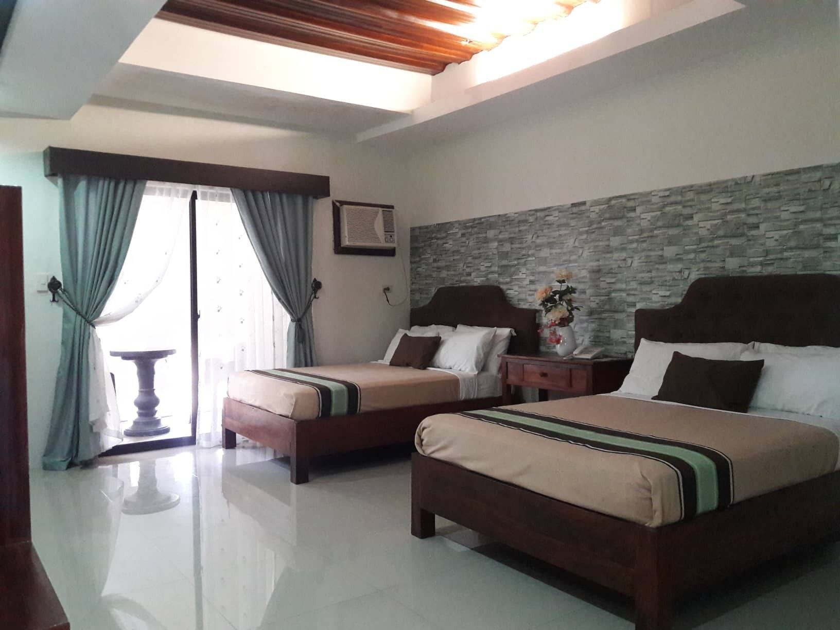 Una habitación de hotel con dos camas, una pared de piedra, una ventana con cortinas y una pequeña mesa con un florero.