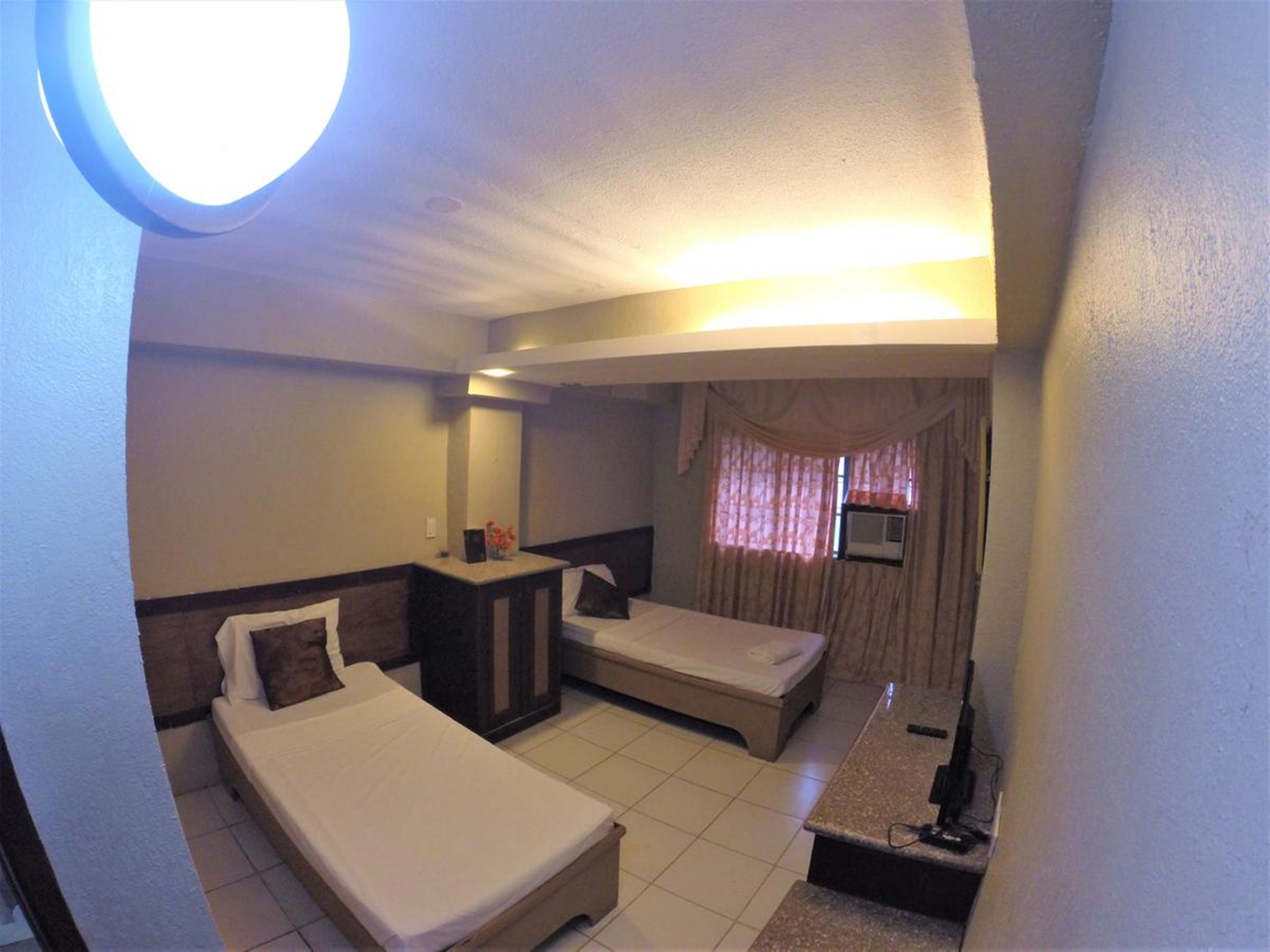 Habitación de hotel con dos camas individuales, un escritorio pequeño, un espejo, una ventana con cortinas y una lámpara en el techo.