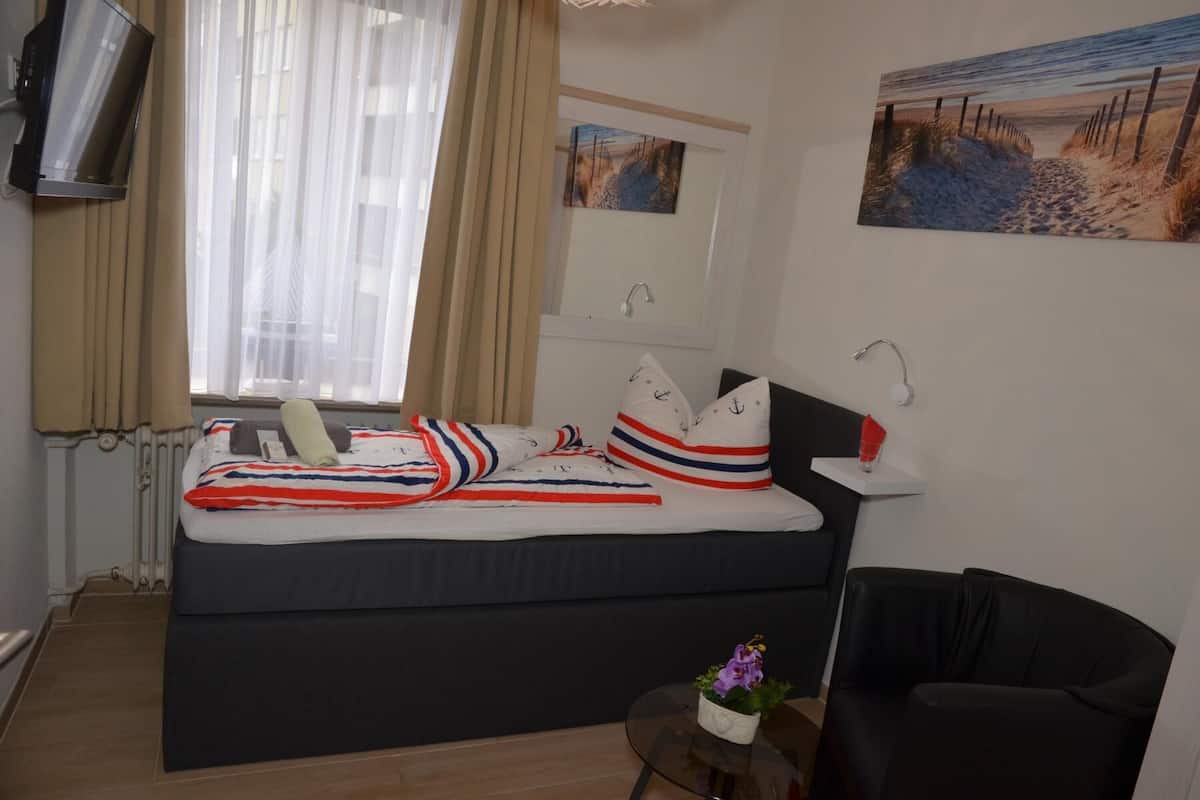 A bedroom with a bed, a small table with a plant, a chair, a window with curtains, a wall-mounted TV, and a painting.