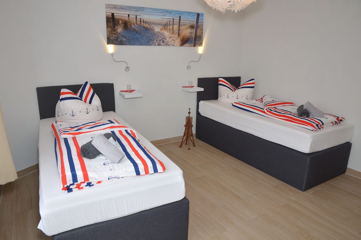 A hotel room with two beds, each with a headboard and a bedside table. There is a painting on the wall above the beds, and a small table with a lamp in the corner.