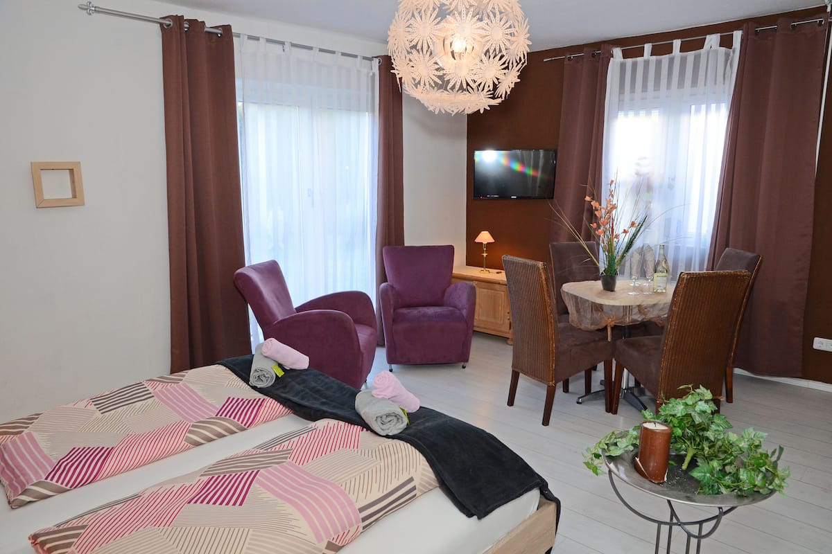 A modern hotel room with a bed, purple armchairs, a dining table with chairs, a TV, and a decorative plant.