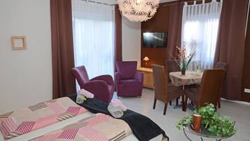 A modern hotel room with a bed, purple armchairs, a dining table with chairs, a TV, and a decorative plant.