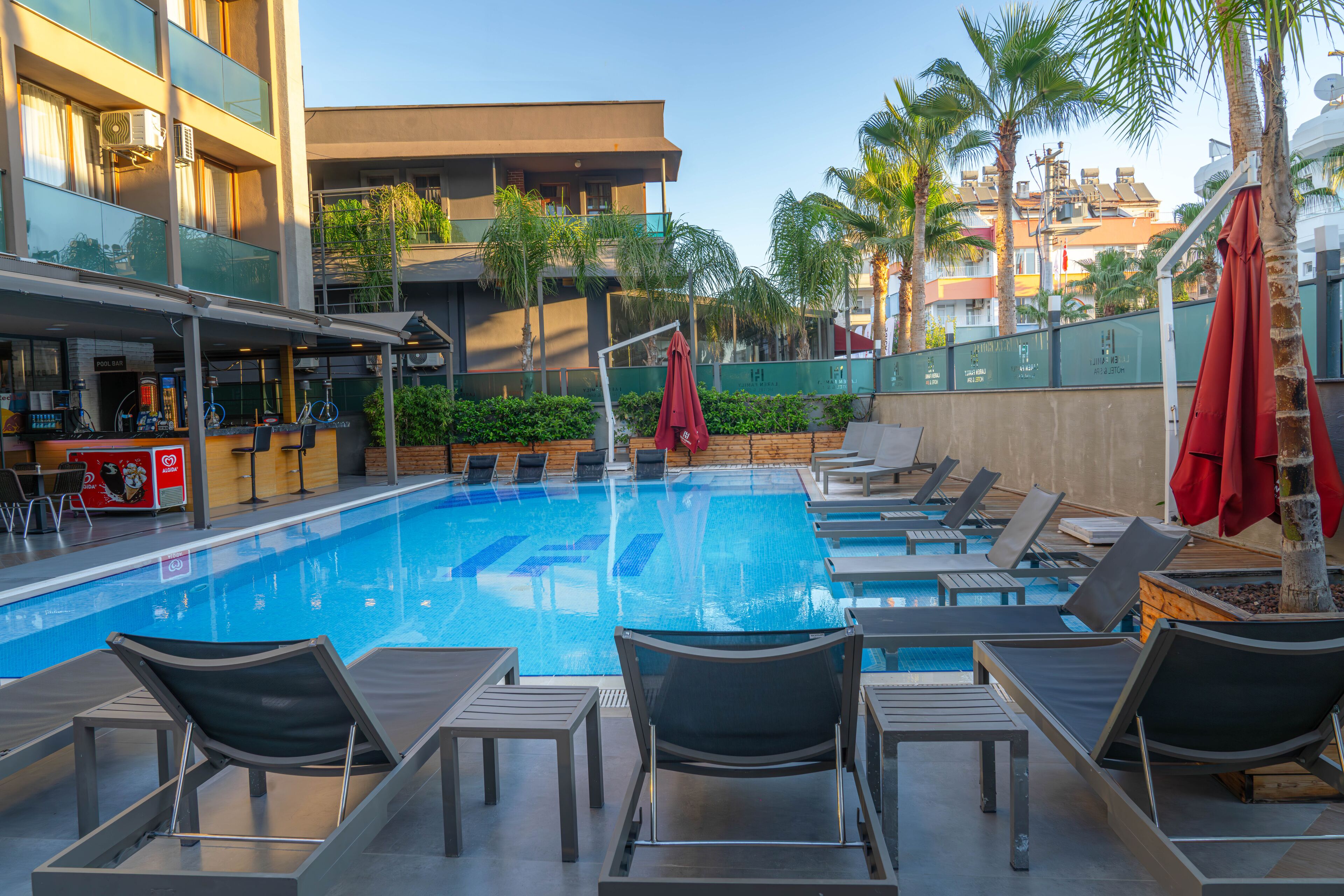 A swimming pool with lounge chairs and umbrellas, surrounded by palm trees and buildings.