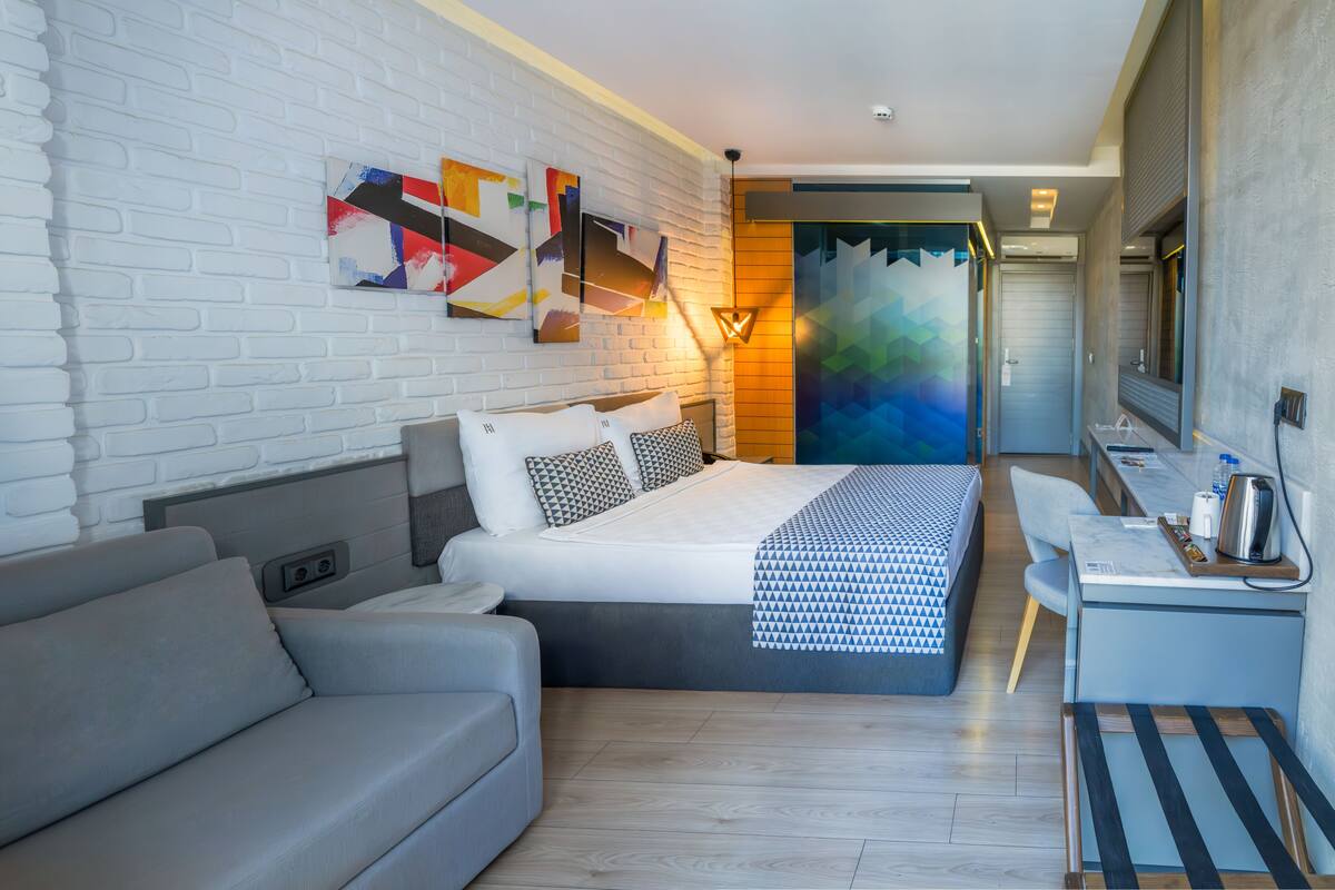 Une chambre d’hôtel moderne avec un lit, un canapé gris, un bureau avec une chaise et un grand miroir.