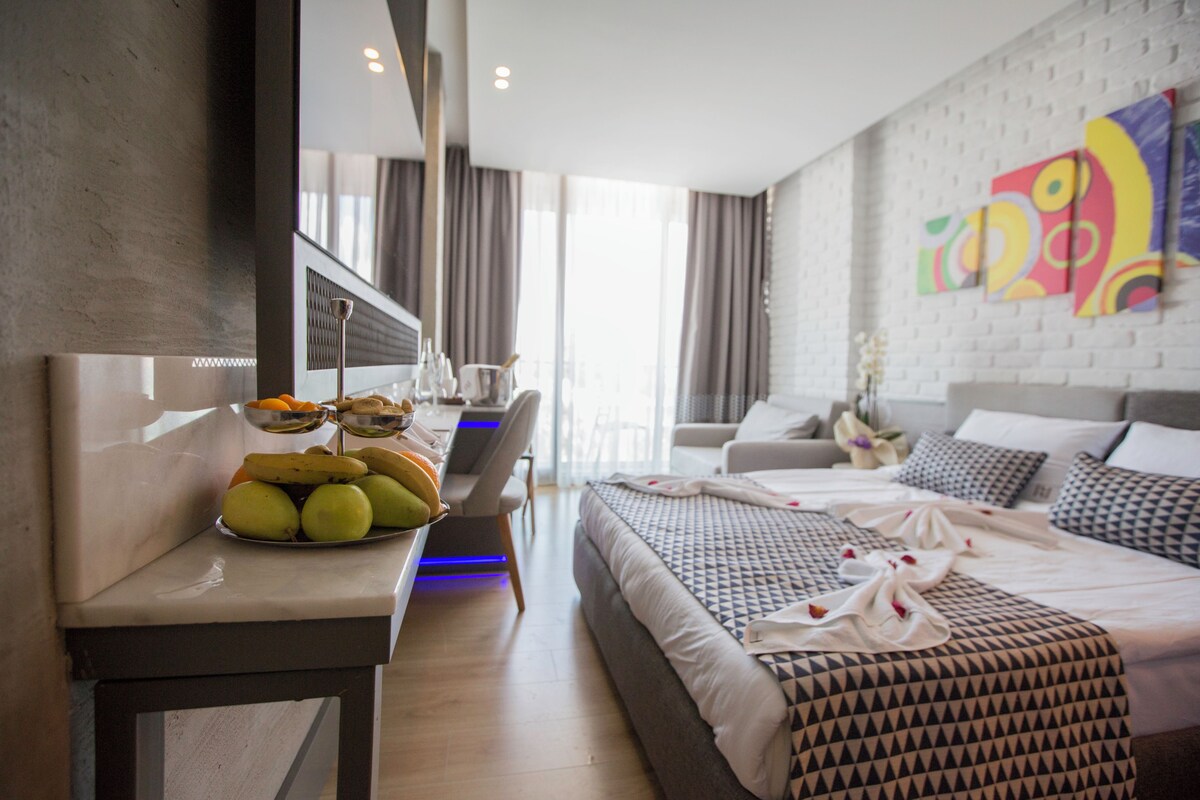 A modern hotel room with a large bed, a bedside table with fruits, a desk, and a view of the city.