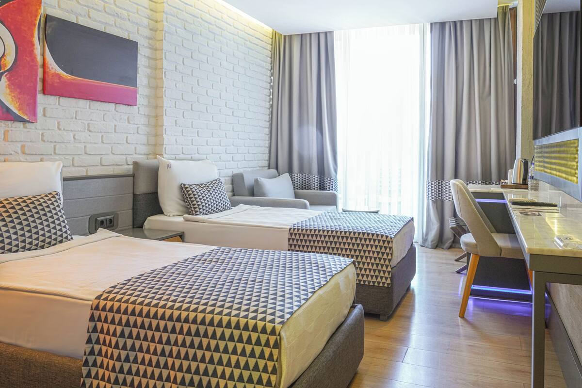 Une chambre d’hôtel avec deux lits, un bureau, une chaise, une grande fenêtre avec des rideaux, un mur en briques blanches et une œuvre d’art colorée.