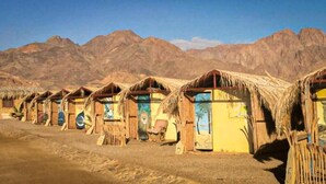A row of thatched-roof huts with simple wooden structures, set against a mountainous backdrop. - Sahara Beach Camp (Nuweiba)