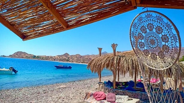 A beach with a thatched roof structure, a dreamcatcher, and a wooden table with cushions. - Sahara Beach Camp (Nuweiba)
