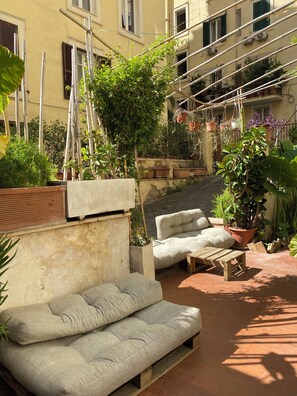 Área externa com assentos, incluindo um sofá e uma mesa pequena, rodeada por plantas em vasos e um edifício com várias janelas e varandas.
