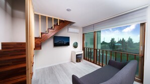 A modern room with a wooden staircase, a grey sofa, a small white desk, and a large window offering a view of trees and buildings. - Poeta Pension (Seogwipo)