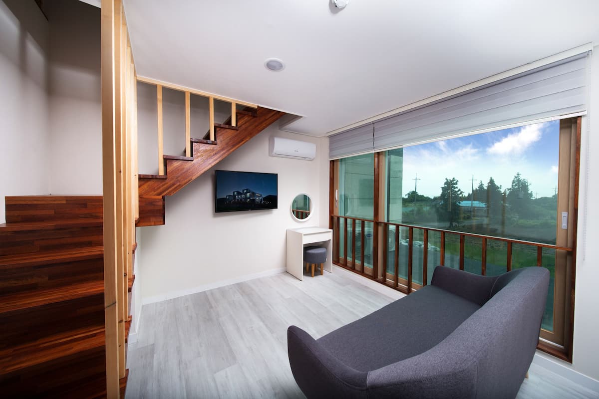 A modern room with a wooden staircase, a grey sofa, a small white desk, and a large window offering a view of trees and buildings.