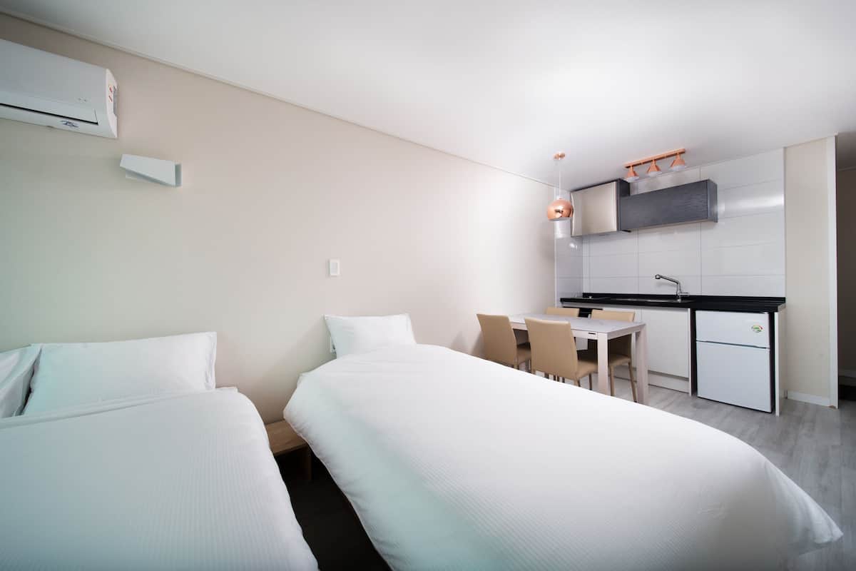 A modern hotel room with two beds, a small kitchenette, and a dining area.