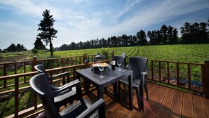 An outdoor deck with a table set for dining, surrounded by chairs, overlooking a grassy field and trees. - Poeta Pension (Seogwipo)